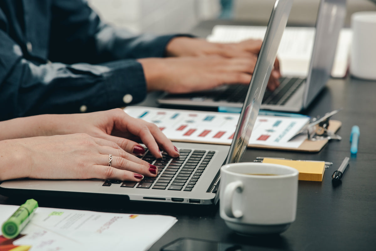 Two people typing on laptops beside coffee and printed charts at a desk
